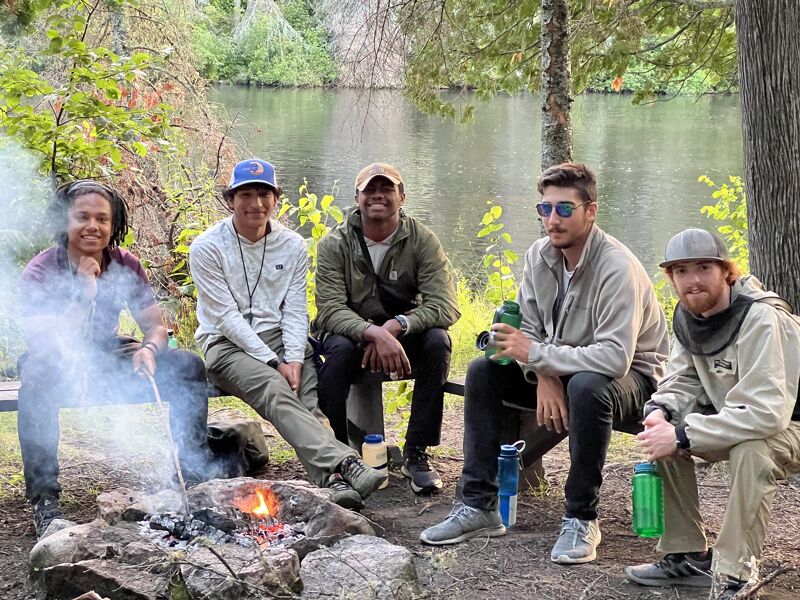 The image shows five young adults gathered around a small campfire near a body of water. They are casually dressed, some wearing hats, and appear to be enjoying a relaxed outdoor setting. The scene suggests a camping trip or a casual gathering in nature, with trees and foliage visible in the background.